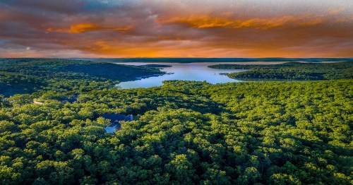 Aerial view of a lush green landscape with a serene lake under a colorful sunset sky.