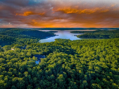Aerial view of a lush green landscape with a winding lake under a dramatic orange and blue sky at sunset.