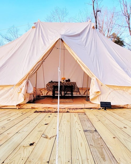 A spacious canvas tent with a wooden deck, featuring a table set inside and surrounded by trees under a clear sky.
