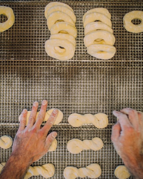 Hands shaping dough into bagels on a textured surface, with various dough shapes arranged nearby.