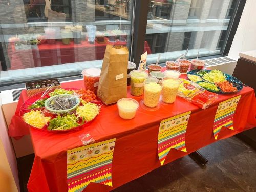 A colorful table with a red tablecloth, featuring various toppings and ingredients for a taco bar, near a window.