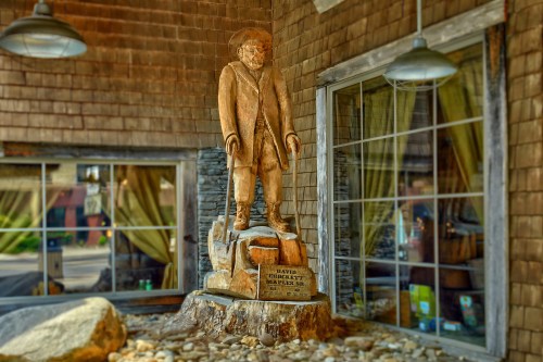 A wooden statue of a man in a hat and coat stands on a stone base, displayed near large windows.