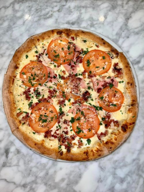 A round pizza topped with sliced tomatoes, bacon, and herbs on a marble surface.