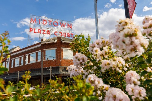 Midtown Plaza Court sign atop a brick building, framed by blooming flowers and a blue sky with clouds.