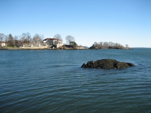 A calm waterfront scene featuring a rocky outcrop and a distant house surrounded by trees under a clear blue sky.