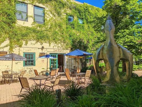 A sunny outdoor patio with tables and chairs, surrounded by greenery and a large abstract sculpture.