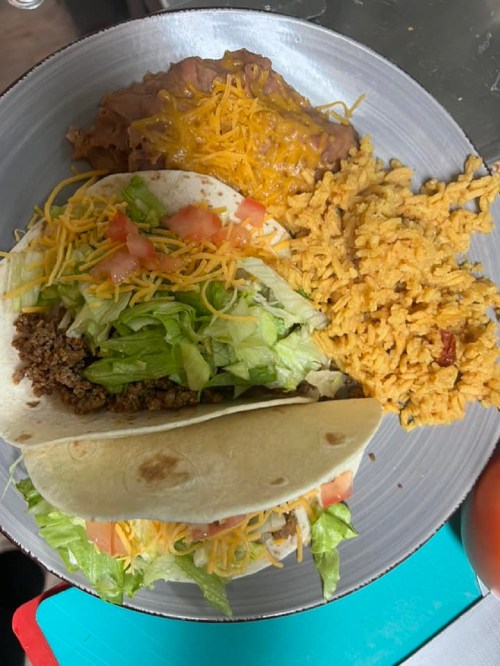 A plate with two tacos filled with ground beef, lettuce, tomatoes, cheese, refried beans, and seasoned rice.