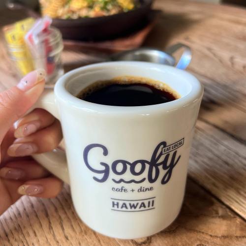A hand holding a white coffee mug with "Goofy Eat Local" printed on it, filled with dark coffee.