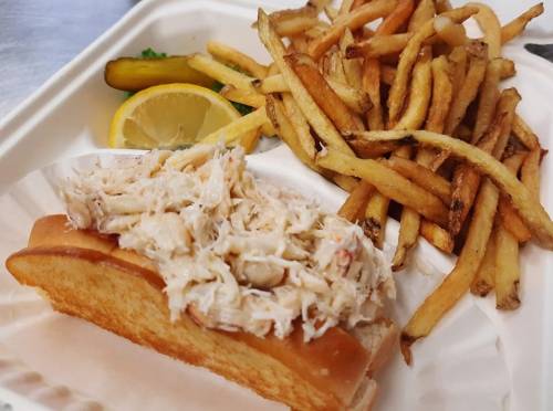 A plate with a crab roll topped with crab meat, served with fries and a lemon wedge.