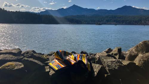 A colorful bag rests on rocks by a serene lake, with mountains and a clear blue sky in the background.