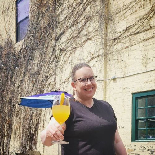 A person holding a cocktail smiles in front of a brick wall with vines and a blue umbrella in the background.