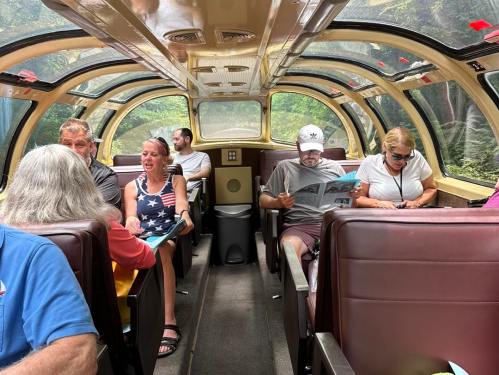 Interior of a train car with passengers reading and chatting, featuring large windows and a cozy atmosphere.
