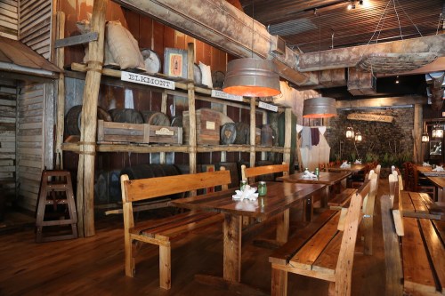 Rustic restaurant interior with wooden tables, benches, and shelves displaying barrels and decor. Warm, inviting atmosphere.