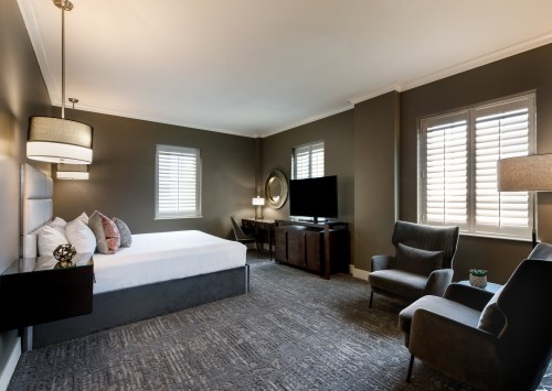 A modern hotel room featuring a bed, armchairs, a TV, and large windows with shutters.