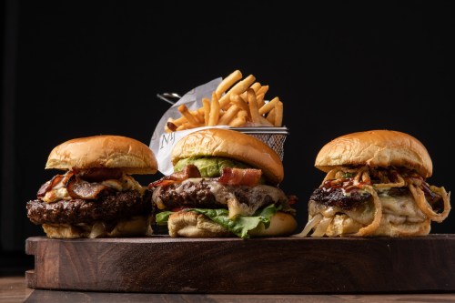 Three gourmet burgers with toppings on a wooden board, accompanied by a basket of golden French fries.