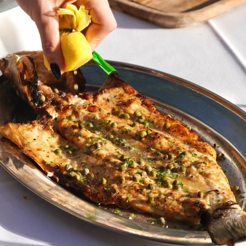 A hand squeezes lemon over a grilled fish garnished with herbs on a silver platter.