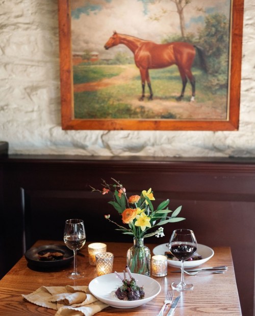 A cozy restaurant table with two plates, wine glasses, a flower vase, and a horse painting on the wall.