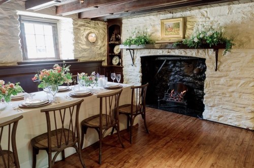 A cozy dining room with a long table set for a meal, a stone fireplace, and floral decorations on the mantel.