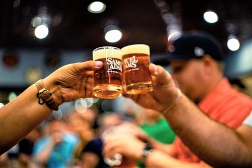 Two hands clink glasses of beer in a lively bar setting, with a blurred crowd in the background.