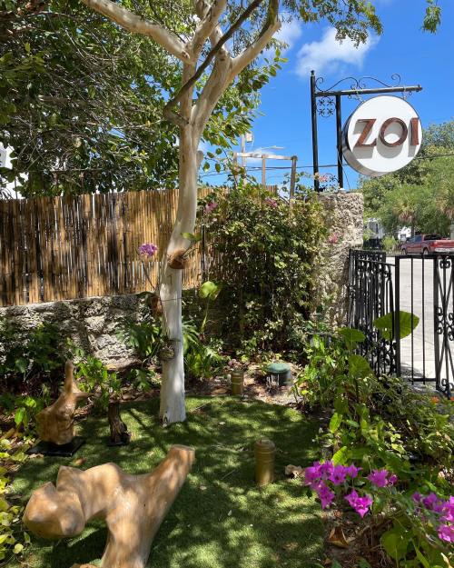A lush garden with greenery and purple flowers, featuring a sign that reads "Zoi" against a clear blue sky.