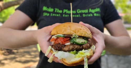 A person holding a juicy burger with lettuce, tomato, jalapeños, and a toothpick, wearing a shirt with a green slogan.
