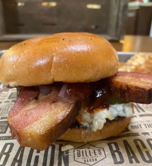 A close-up of a bacon sandwich with a glossy bun, crispy bacon, and toppings on a paper-lined tray.