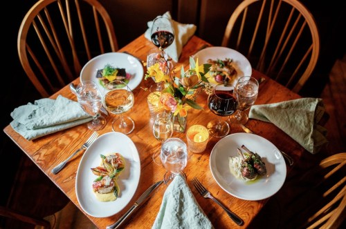 A beautifully set dining table with four plates of gourmet food, glasses of wine, and a floral centerpiece.