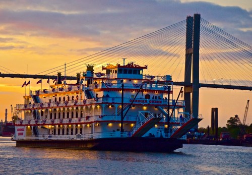 A steamboat named "Georgia Queen" sails under a bridge at sunset, with vibrant colors reflecting on the water.