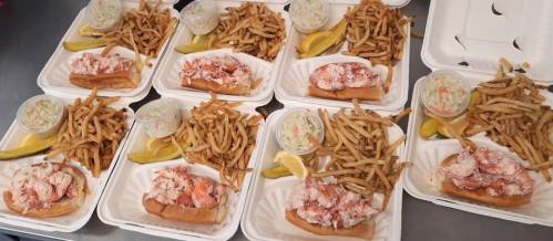 Six takeout trays with lobster rolls, fries, coleslaw, and lemon wedges arranged on a table.