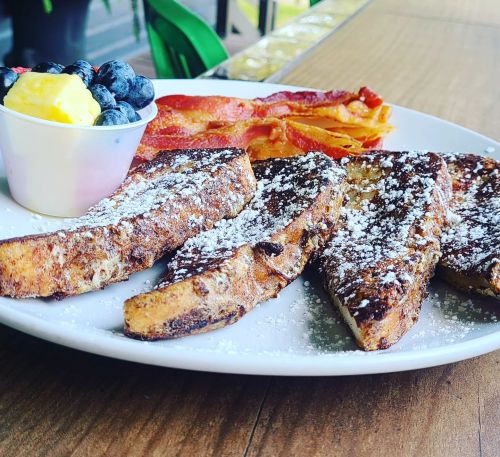 A plate of French toast dusted with powdered sugar, served with bacon and a side of fruit.