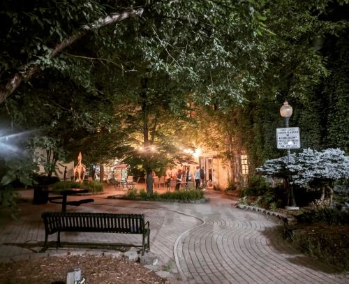 A dimly lit park path leads to a cozy gathering area surrounded by trees and greenery.