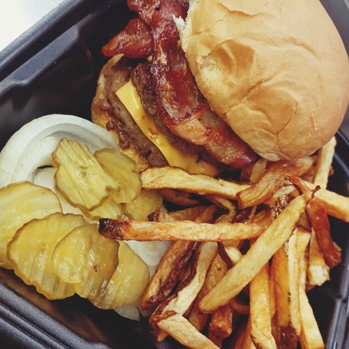 A close-up of a burger with bacon and cheese, served with pickles, onions, and crispy French fries in a takeout container.