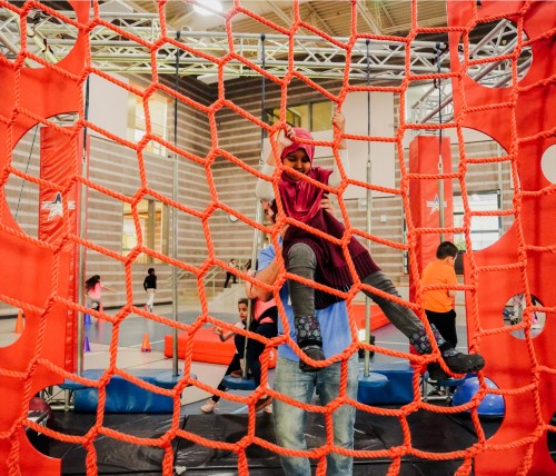 A child climbs a red net structure in a gym, while an adult supports them from below. Other kids play in the background.