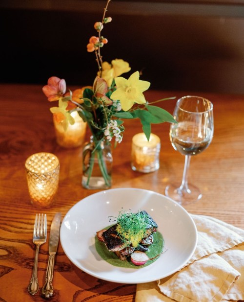 A beautifully plated dish with greens, garnished with herbs, beside a glass of wine and a floral centerpiece on a wooden table.