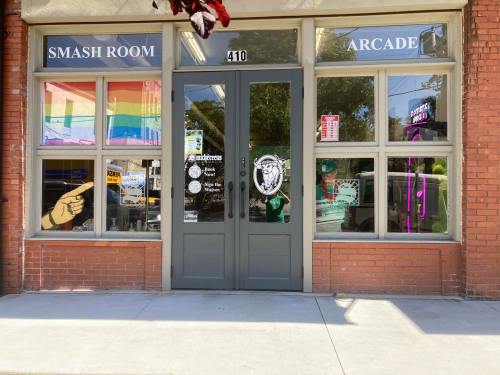 Exterior of a storefront featuring "Smash Room" and "Arcade" signs, with rainbow flags and various colorful window decals.
