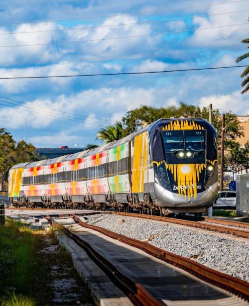 A colorful train approaches on tracks, surrounded by greenery and under a partly cloudy sky.