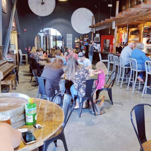 A lively bar with patrons seated at tables and the bar, enjoying drinks and food in a spacious, rustic setting.