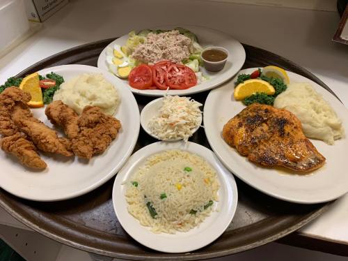 A tray with two plates of fried chicken, grilled fish, rice, coleslaw, and fresh vegetables.