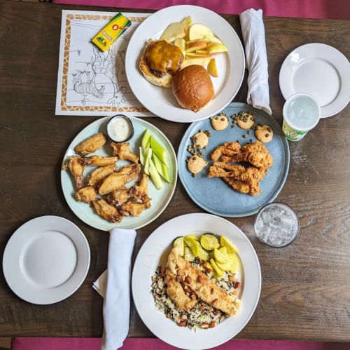 A table set with plates of chicken wings, a burger, fish with rice, apple slices, cookies, and drinks.