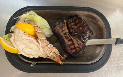 A metal tray holds a grilled steak and a lobster tail, garnished with lemon and lettuce.