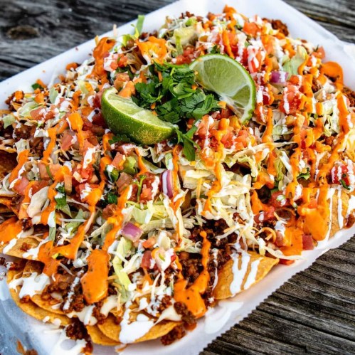 A plate of loaded nachos topped with cheese, lettuce, tomatoes, onions, cilantro, and lime wedges.