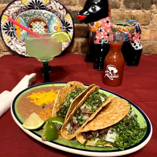 Three tacos filled with meat and topped with cilantro, served with lime, rice, beans, and a margarita in the background.