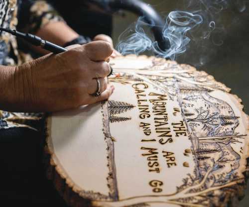 A person woodburns a sign reading "The Mountains Are Calling and I Must Go," with smoke rising from the tool.