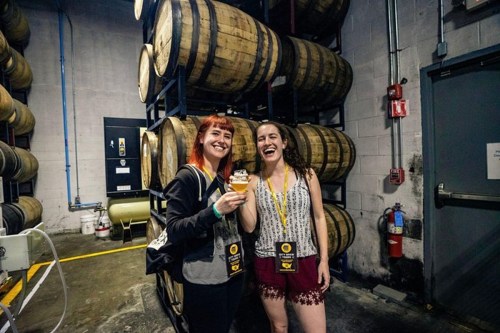 Two women smile and hold glasses of beer in a brewery with stacked barrels in the background.