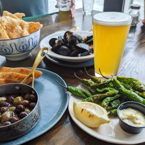 A table with various dishes: olives, fried snacks, mussels, grilled peppers, and a glass of beer.