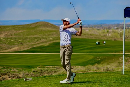 A golfer swings a club on a lush green course with rolling hills in the background under a clear blue sky.