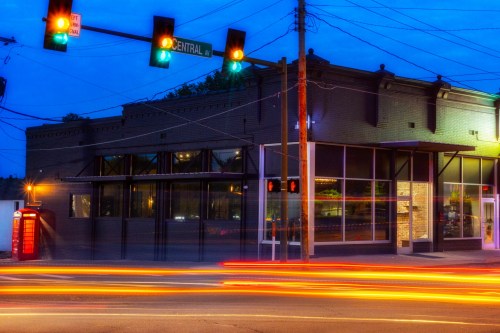 A street view of a building at dusk, with traffic lights and light trails from passing cars.