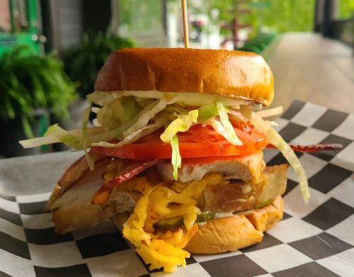 A stacked sandwich with layers of meat, cheese, lettuce, tomato, and pickles on a toasted bun, served on checkered paper.