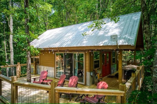 A cozy cabin nestled in a forest, featuring a deck with red chairs and large windows surrounded by greenery.
