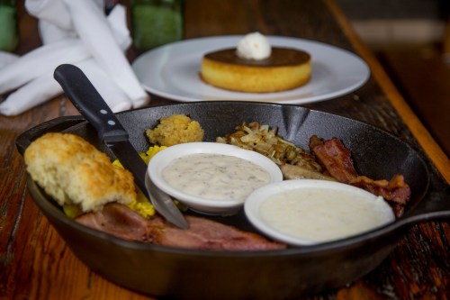 A cast iron skillet filled with bacon, sausage, biscuits, gravy, and hash browns, with a dessert in the background.